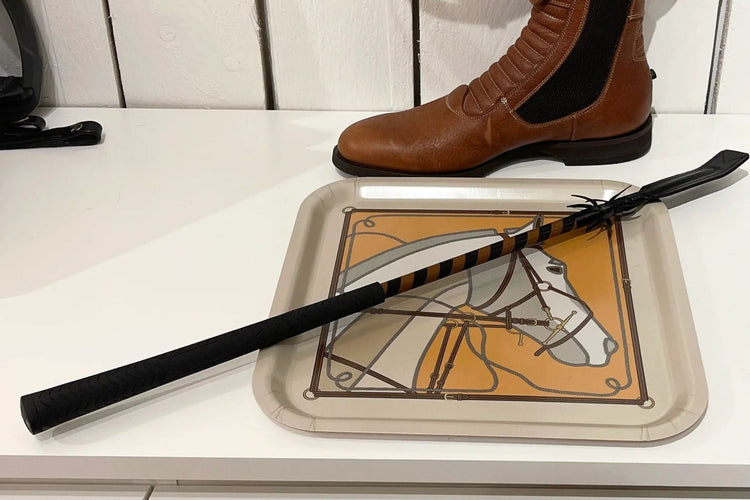 Cravache d’équitation noire avec poignée antidérapante et claquette plate, posée sur un plateau décoré cheval, accessoire pour obstacle et entraînement
